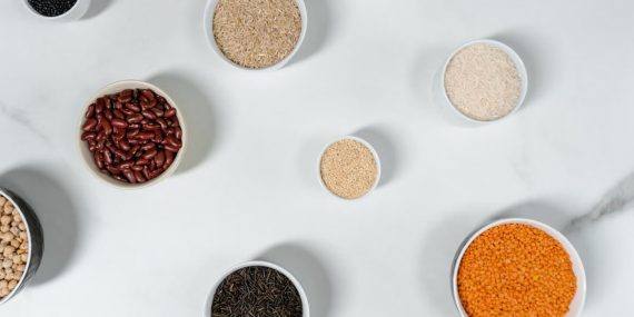 Top view of various grains and legumes in bowls on a white marble surface.